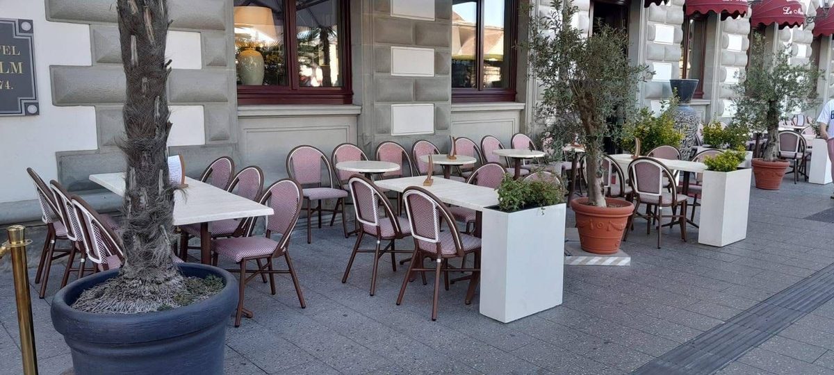 Dehors di un locale con tavoli in marmo chiaro, sedute intrecciate con struttura marrone, ulivi e fioriere bianche
