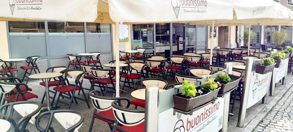 Dehors di una gelateria con ombrelloni, tavolini rotondi e sedie pieghevoli con cuscini rossi, fioriere sul bordo